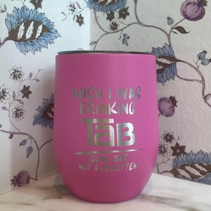 Op de afbeelding: Een felroze ge&iuml;soleerde tumbler met zilverkleurige tekst "WISH I WAS DRINKING TAB GONE BUT NOT FORGOTTEN". De tumbler heeft een afgeronde vorm en een zwart deksel. De achtergrond toont bloemenbehang.