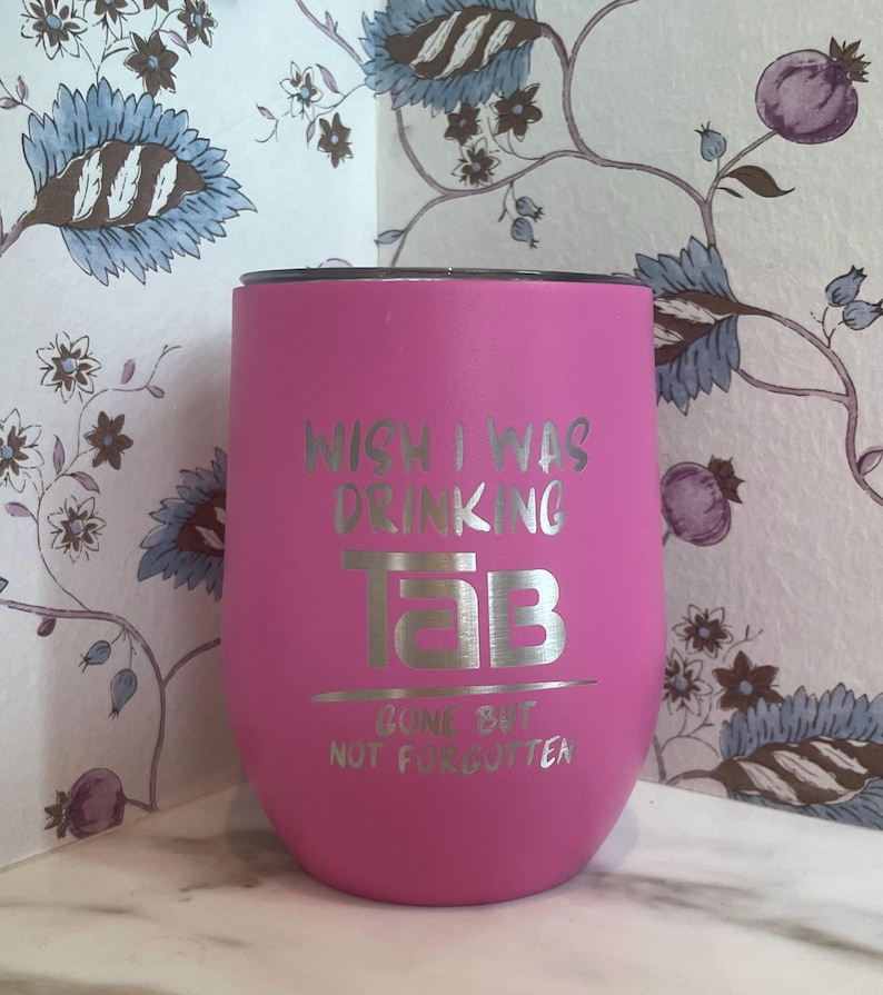 Op de afbeelding: Een felroze ge&iuml;soleerde tumbler met zilverkleurige tekst "WISH I WAS DRINKING TAB GONE BUT NOT FORGOTTEN". De tumbler heeft een afgeronde vorm en een zwart deksel. De achtergrond toont bloemenbehang.