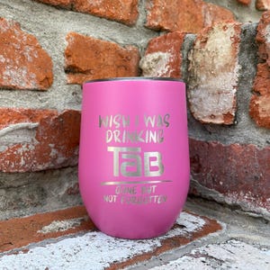 Op de afbeelding: Een roze ge&iuml;soleerde tumbler met de tekst "Wish I Was Drinking Tab" en "Gone But Not Forgotten" in zilveren letters. De tumbler is cilindrisch met een afgeronde basis en een zwart deksel. De achtergrond is een bakstenen muur.