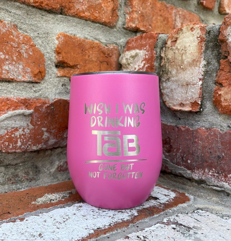 Op de afbeelding: Een roze ge&iuml;soleerde tumbler met de tekst "Wish I Was Drinking Tab" en "Gone But Not Forgotten" in zilveren letters. De tumbler is cilindrisch met een afgeronde basis en een zwart deksel. De achtergrond is een bakstenen muur.