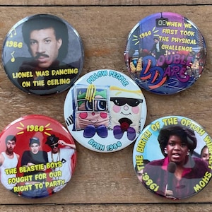 May include: A collection of five round buttons with various designs and text. One button features Lionel Richie, another has the text "Double Dare", and a third displays The Beastie Boys. Other buttons show Oprah Winfrey and a Pillow People character.