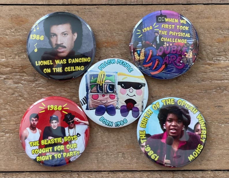 May include: A collection of five round buttons with various designs and text. One button features Lionel Richie, another has the text "Double Dare", and a third displays The Beastie Boys. Other buttons show Oprah Winfrey and a Pillow People character.
