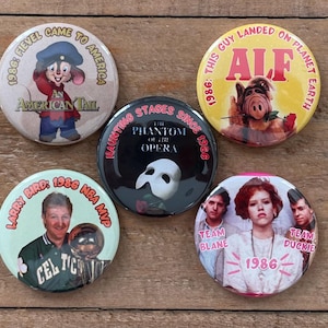 May include: A collection of five round pinback buttons. One features Fievel from "An American Tail," another ALF, a third Larry Bird, and a fourth with the text "Team Blane" and "Team Duckie." The fifth button has a Phantom of the Opera design.