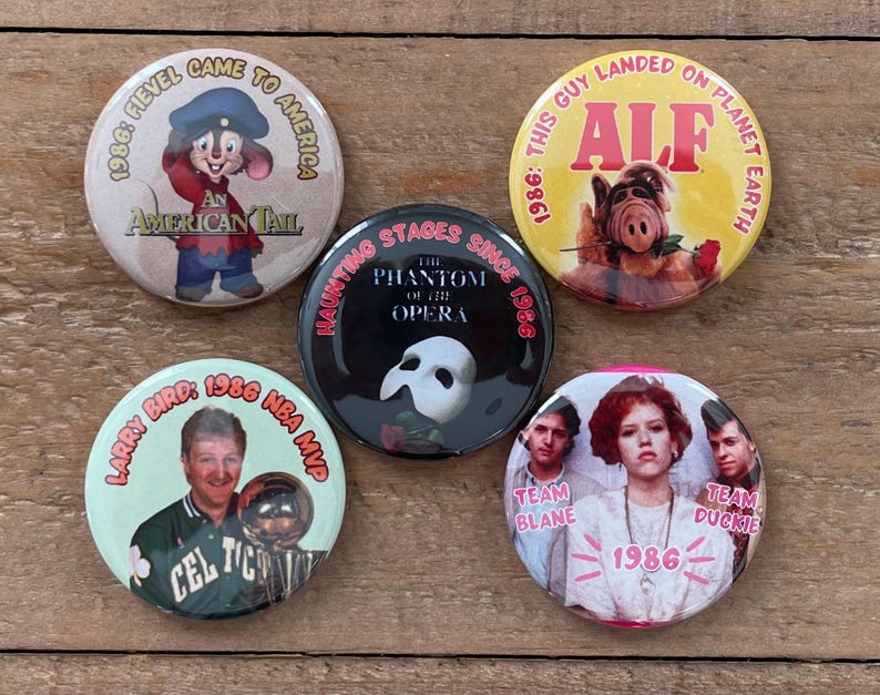 May include: A collection of five round pinback buttons. One features Fievel from "An American Tail," another ALF, a third Larry Bird, and a fourth with the text "Team Blane" and "Team Duckie." The fifth button has a Phantom of the Opera design.