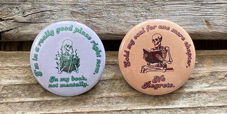 Pode incluir: Dois bot&otilde;es redondos com ilustra&ccedil;&otilde;es de esqueletos. Um &eacute; lil&aacute;s claro com o texto: "I'm in a really good place right now. In my book, not mentally." O outro &eacute; p&ecirc;ssego com o texto: "Sold my soul for one more chapter. No Regrets."
