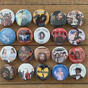 May include: A collection of twenty-one round pinback buttons featuring album art and portraits of musicians. The buttons showcase a variety of colors and designs, with text and images of artists and album titles.