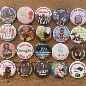 May include: A collection of colorful, round pinback buttons with various pop culture references from the 1990s. The buttons feature images of celebrities, movies, and catchphrases, with text and graphics. The buttons are arranged on a wooden surface.