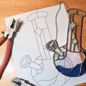 Könnte beinhalten: Eine Glasbong mit blauem, lila und klarem Design hängt an einer silbernen Kette. Eine Strichzeichnung einer Bong befindet sich auf einem weißen Blatt Papier. Zwei Zangen mit roten Griffen liegen auf einer Holzoberfläche.