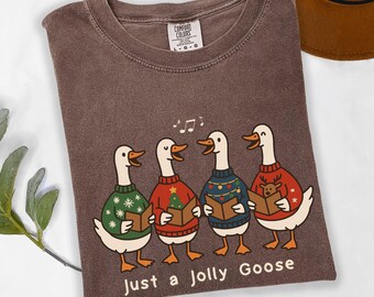 May include: Brown t-shirt featuring a graphic of four geese wearing festive sweaters and singing from songbooks. The text "Just a Jolly Goose" is printed below the geese. A hat is visible in the upper right corner.