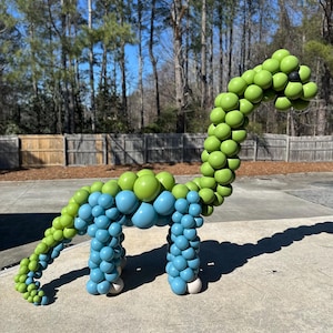 Puede incluir: Una escultura de dinosaurio hecha con globos verdes y azules. El dinosaurio tiene un cuello y cola largos, con pies blancos. La escultura está al aire libre en un día soleado.