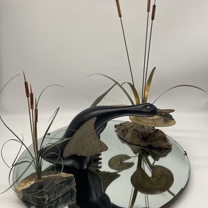 Puede incluir: Una escultura de pato negro con las alas extendidas, sosteniendo una rana verde en su pico, est&aacute; posada sobre un espejo circular. El pato se refleja en el espejo, junto con un grupo de juncos marrones y un trozo de madera flotante.