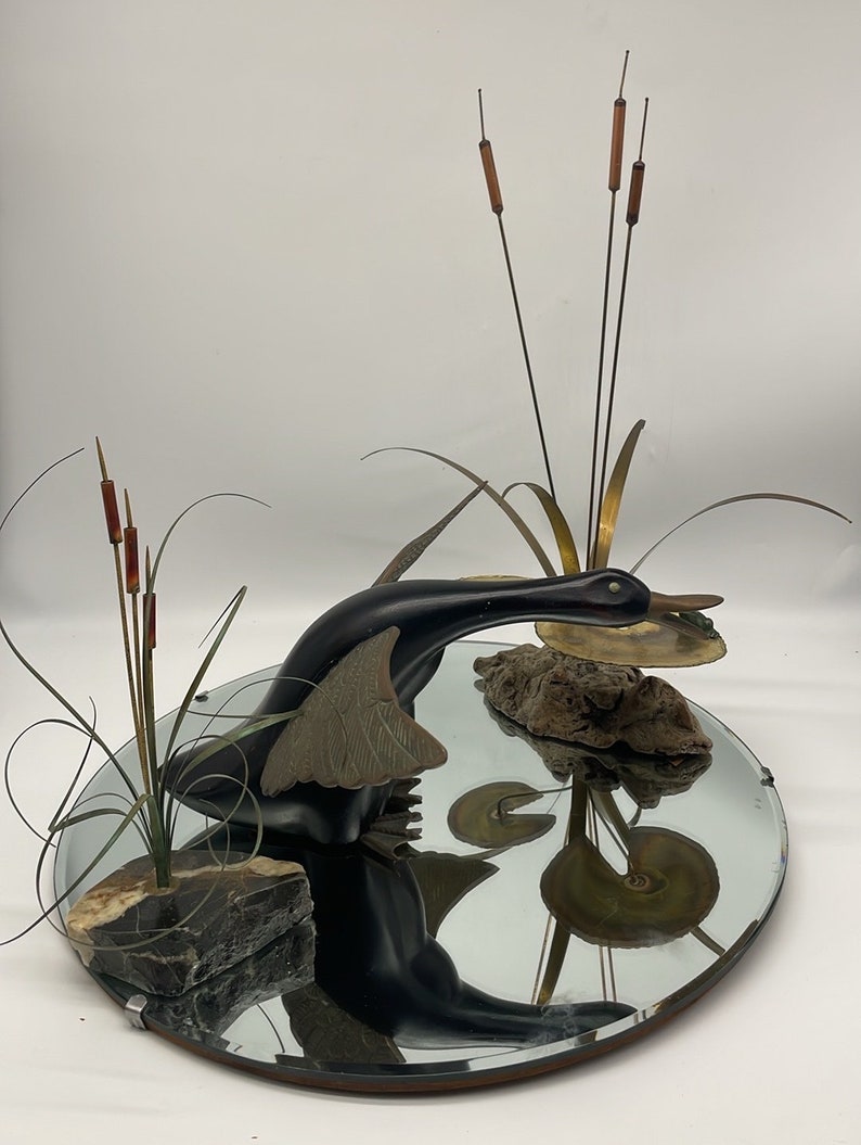 Puede incluir: Una escultura de pato negro con las alas extendidas, sosteniendo una rana verde en su pico, est&aacute; posada sobre un espejo circular. El pato se refleja en el espejo, junto con un grupo de juncos marrones y un trozo de madera flotante.