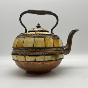 May include: A copper teapot with a bone inlay design and a brass handle. The teapot has a round body and a spout on the side.