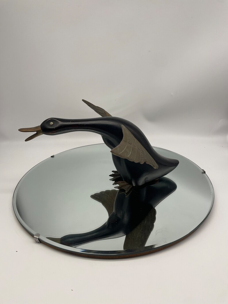 Puede incluir: Una escultura de ganso de madera negra con alas doradas, de pie sobre un espejo redondo con marco plateado.