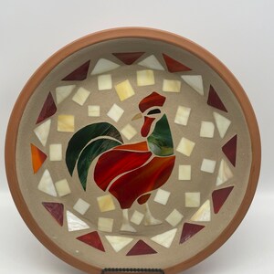 May include: A terracotta ceramic plate with a mosaic design of a rooster. The rooster is made of red, green, and white glass tiles. The plate is decorated with a border of red and white triangles.