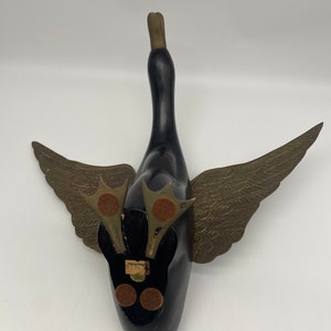Puede incluir: Un ganso de madera negro con alas y cabeza doradas. El ganso est&aacute; acostado de espaldas con las alas extendidas. El ganso tiene una etiqueta en la parte inferior que dice "Woodcarvings 92801".