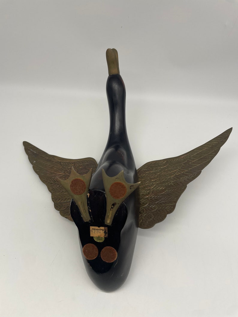 Puede incluir: Un ganso de madera negro con alas y cabeza doradas. El ganso est&aacute; acostado de espaldas con las alas extendidas. El ganso tiene una etiqueta en la parte inferior que dice "Woodcarvings 92801".