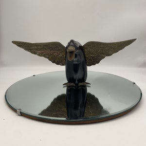 Puede incluir: Una escultura de p&aacute;jaro de madera negra con alas doradas, de pie sobre un espejo redondo con marco de madera.