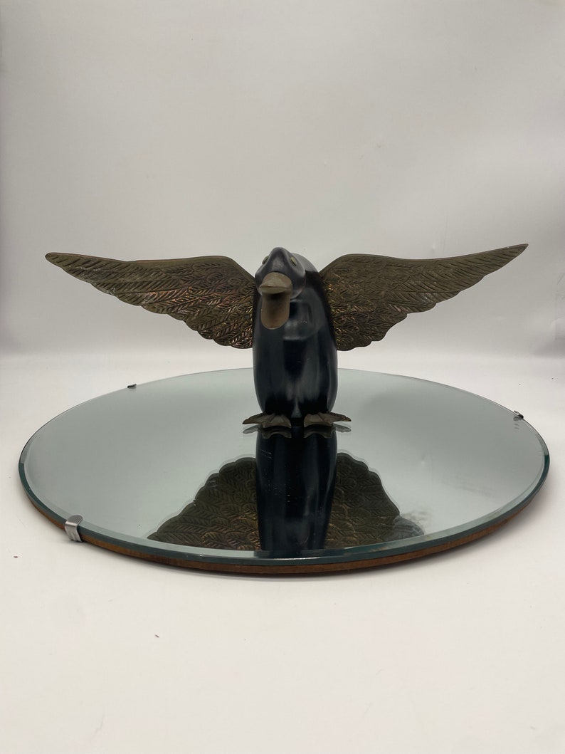 Puede incluir: Una escultura de p&aacute;jaro de madera negra con alas doradas, de pie sobre un espejo redondo con marco de madera.