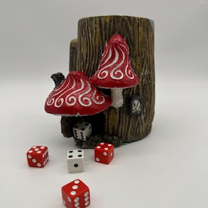 May include: A ceramic mug shaped like a tree stump with two red mushroom houses on top. The mug is designed to hold dice, and there are four dice scattered around the mug, three red and one white.