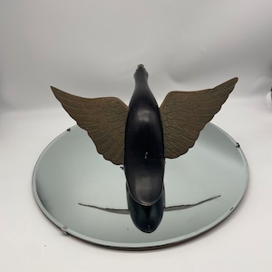Puede incluir: Una escultura de p&aacute;jaro de madera negra con alas doradas. El p&aacute;jaro est&aacute; en posici&oacute;n de vuelo con las alas extendidas. La escultura est&aacute; sentada sobre un espejo redondo plateado.