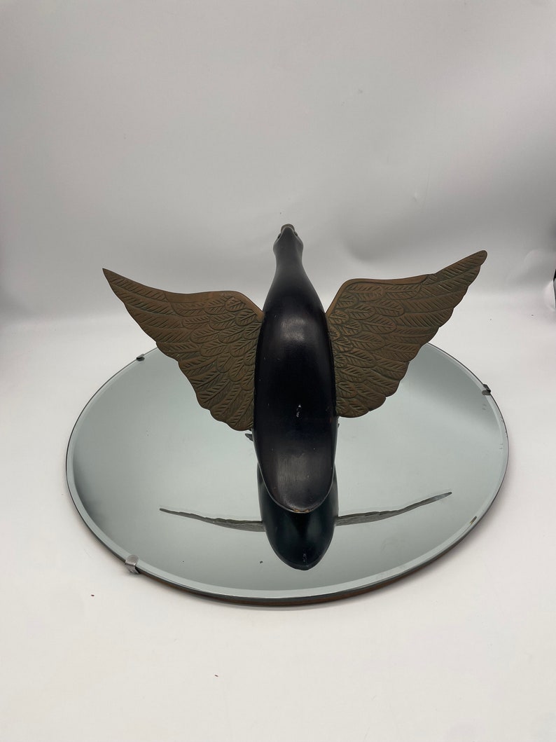 Puede incluir: Una escultura de p&aacute;jaro de madera negra con alas doradas. El p&aacute;jaro est&aacute; en posici&oacute;n de vuelo con las alas extendidas. La escultura est&aacute; sentada sobre un espejo redondo plateado.