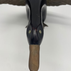 Puede incluir: Un se&ntilde;uelo de pato de madera con plumas pintadas de negro y un pico de lat&oacute;n. El pato tiene ojos grandes y redondos.