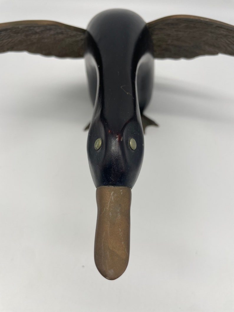 Puede incluir: Un se&ntilde;uelo de pato de madera con plumas pintadas de negro y un pico de lat&oacute;n. El pato tiene ojos grandes y redondos.