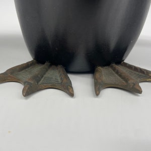 Puede incluir: Primer plano de la parte inferior de una figurita de ping&uuml;ino de cer&aacute;mica negra. El ping&uuml;ino tiene pies de tres dedos hechos de metal con un acabado bronce.