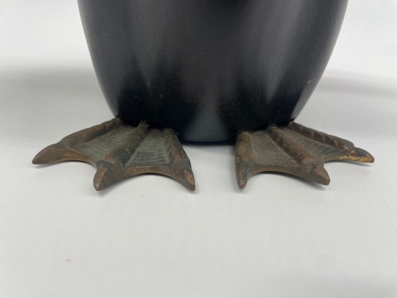Puede incluir: Primer plano de la parte inferior de una figurita de ping&uuml;ino de cer&aacute;mica negra. El ping&uuml;ino tiene pies de tres dedos hechos de metal con un acabado bronce.