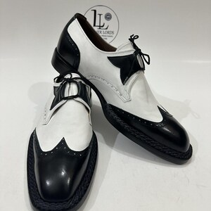 May include: A pair of black and white wingtip oxford shoes featuring a two-tone leather design, black laces, and a braided trim detail along the sole edge. The shoes have a polished finish with decorative perforations on the toe cap.