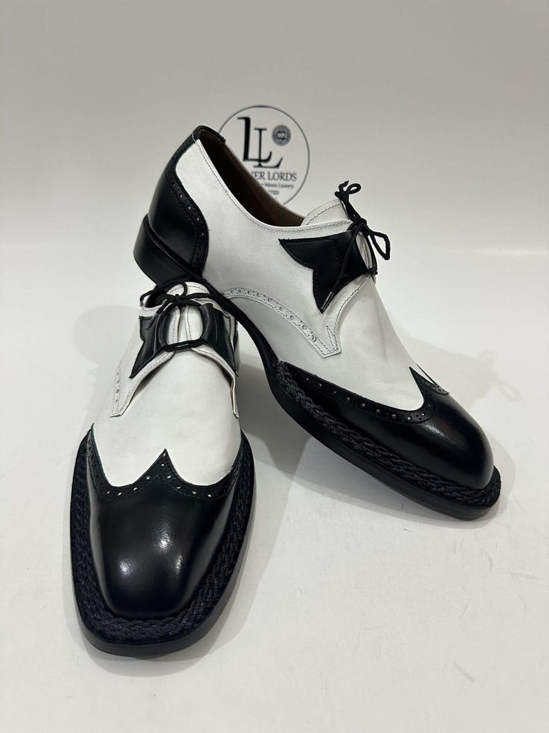 May include: A pair of black and white wingtip oxford shoes featuring a two-tone leather design, black laces, and a braided trim detail along the sole edge. The shoes have a polished finish with decorative perforations on the toe cap.