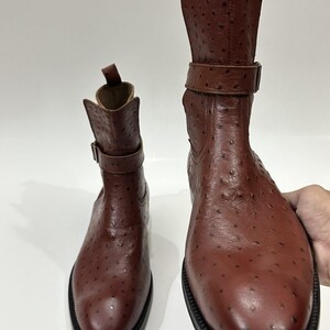 May include: A pair of brown ankle boots featuring a textured ostrich-style leather finish, a rounded toe, a pull tab at the heel, and a decorative strap with a metal buckle around the upper shaft, set on a black sole.