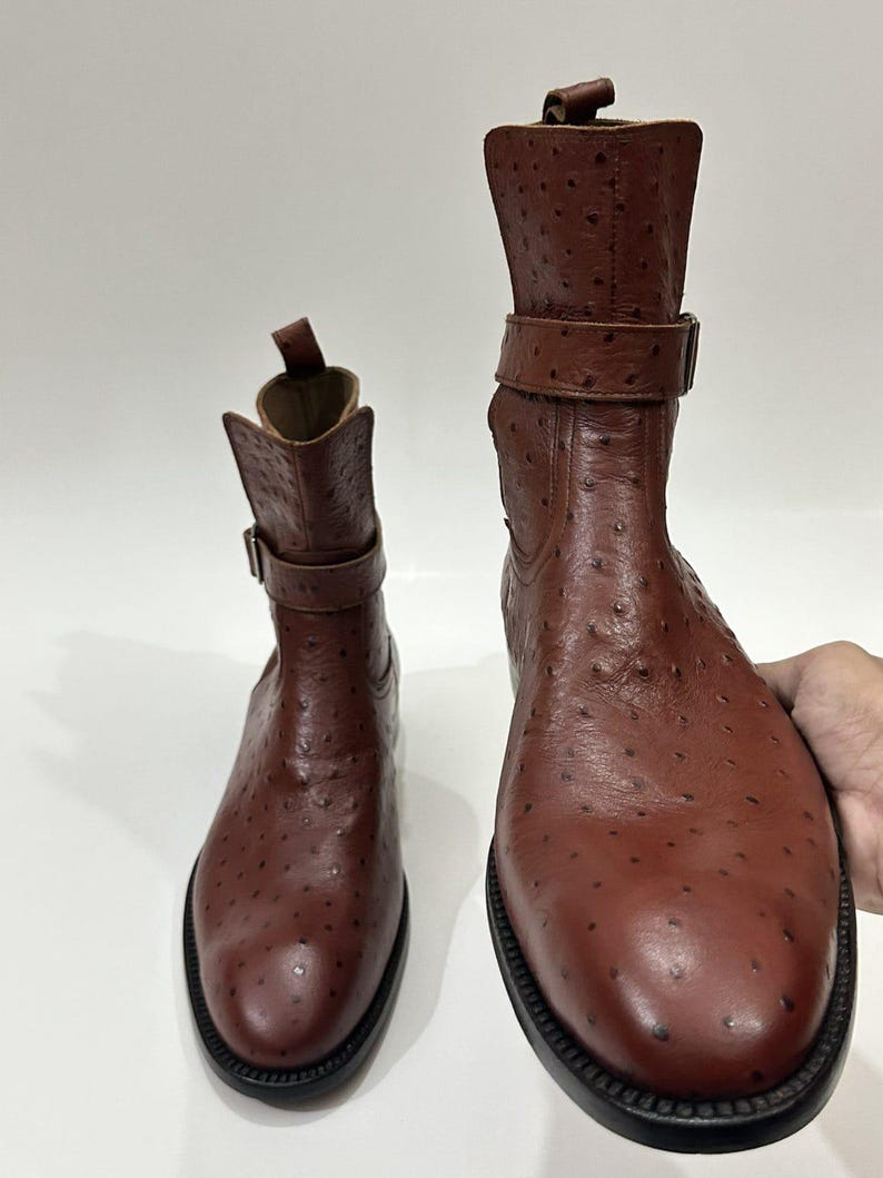 May include: A pair of brown ankle boots featuring a textured ostrich-style leather finish, a rounded toe, a pull tab at the heel, and a decorative strap with a metal buckle around the upper shaft, set on a black sole.
