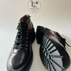 May include: A pair of dark brown leather boots with black laces, brass-toned eyelets, and thick black lug soles. One boot is upright, and the other is positioned on its side to show the tread. A circular logo reads LEATHER LORDS.