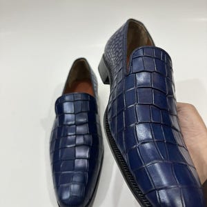 May include: A pair of navy blue loafers featuring a textured crocodile-embossed pattern across the upper. The slip-on shoes have a rounded toe, a low heel, and a smooth tan interior lining, showcasing a structured and polished appearance.