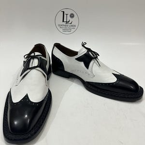 May include: A pair of black and white leather wingtip oxford shoes featuring a braided black welt, black laces, and a logo on the background reading LEATHER LORDS Where Leather Meets Luxury HANDCRAFTED Leather Products.