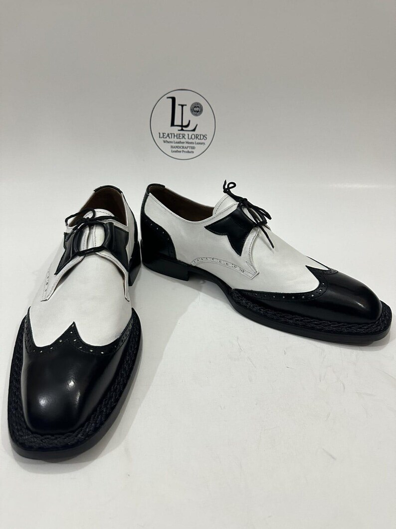 May include: A pair of black and white leather wingtip oxford shoes featuring a braided black welt, black laces, and a logo on the background reading LEATHER LORDS Where Leather Meets Luxury HANDCRAFTED Leather Products.