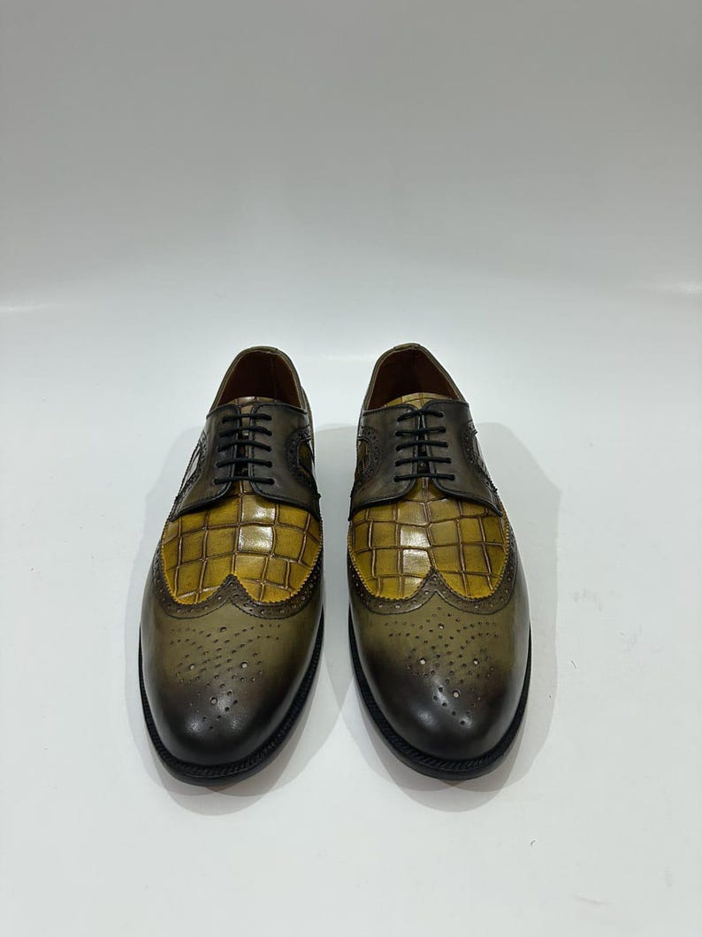 Puede incluir: Un par de zapatos oxford con un dise&ntilde;o bicolor que presenta un empeine de cuero con relieve de cocodrilo en color amarillo mostaza y una puntera y tal&oacute;n de cuero bru&ntilde;ido en verde oliva oscuro. Incluyen cordones negros y detalles brogue.