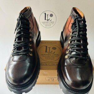 May include: A pair of dark brown leather boots with black laces and brass-toned eyelets. The boots feature a thick black lug sole and a gradient finish. A cardboard box with the text LEATHER LORDS is positioned between the boots.