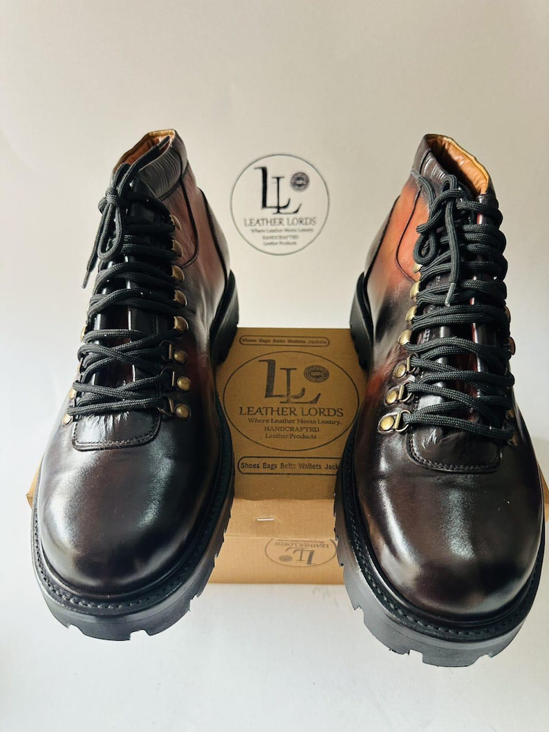 May include: A pair of dark brown leather boots with black laces and brass-toned eyelets. The boots feature a thick black lug sole and a gradient finish. A cardboard box with the text LEATHER LORDS is positioned between the boots.