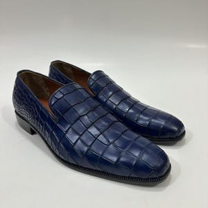 May include: A pair of deep blue slip-on loafers featuring a prominent embossed crocodile skin texture across the entire upper surface. The shoes have a rounded toe, a low heel, and a dark sole, with a smooth tan interior lining visible.