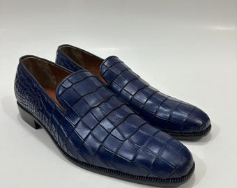 Puede incluir: Un par de mocasines azul oscuro con una textura en relieve de piel de cocodrilo en toda la superficie superior. Los zapatos tienen punta redondeada, tacón bajo y suela oscura, con un forro interior de color tostado visible.