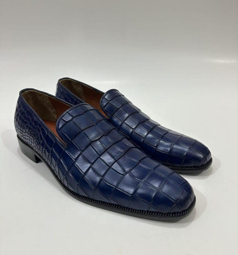 May include: A pair of deep blue slip-on loafers featuring a prominent embossed crocodile skin texture across the entire upper surface. The shoes have a rounded toe, a low heel, and a dark sole, with a smooth tan interior lining visible.