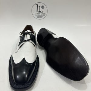 May include: A pair of black and white wingtip oxford shoes with black laces and a braided trim along the sole. One shoe is upright showing the upper, while the other is placed sole-up. A circular logo with the text LEATHER LORDS is visible.
