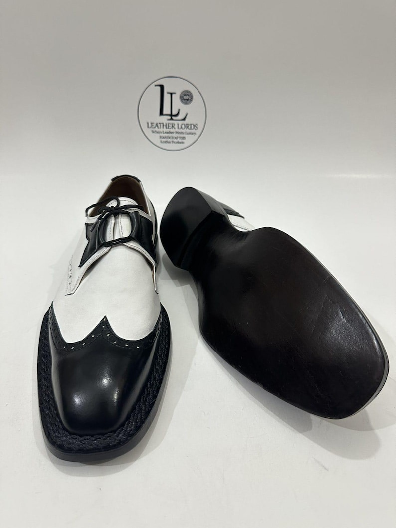 May include: A pair of black and white wingtip oxford shoes with black laces and a braided trim along the sole. One shoe is upright showing the upper, while the other is placed sole-up. A circular logo with the text LEATHER LORDS is visible.