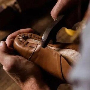 May include: A tan leather shoe being worked on with a metal tool. The shoe features visible dark stitching along the toe area, securing the leather upper to the sole. The material appears smooth and light brown in color.