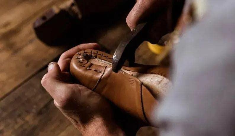 May include: A tan leather shoe being worked on with a metal tool. The shoe features visible dark stitching along the toe area, securing the leather upper to the sole. The material appears smooth and light brown in color.