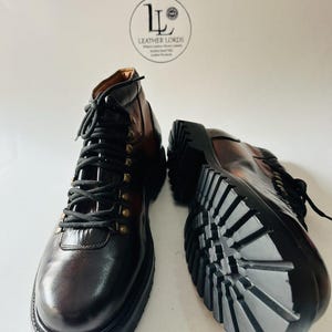 May include: A pair of dark brown leather boots with black laces, brass-toned eyelets, and thick black lug soles. One boot is upright, and the other is positioned on its side to show the tread. A circular logo reads LEATHER LORDS.