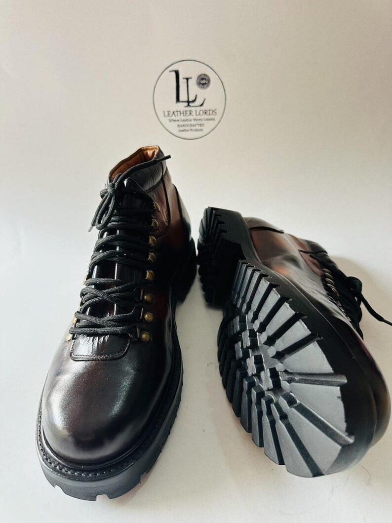 May include: A pair of dark brown leather boots with black laces, brass-toned eyelets, and thick black lug soles. One boot is upright, and the other is positioned on its side to show the tread. A circular logo reads LEATHER LORDS.